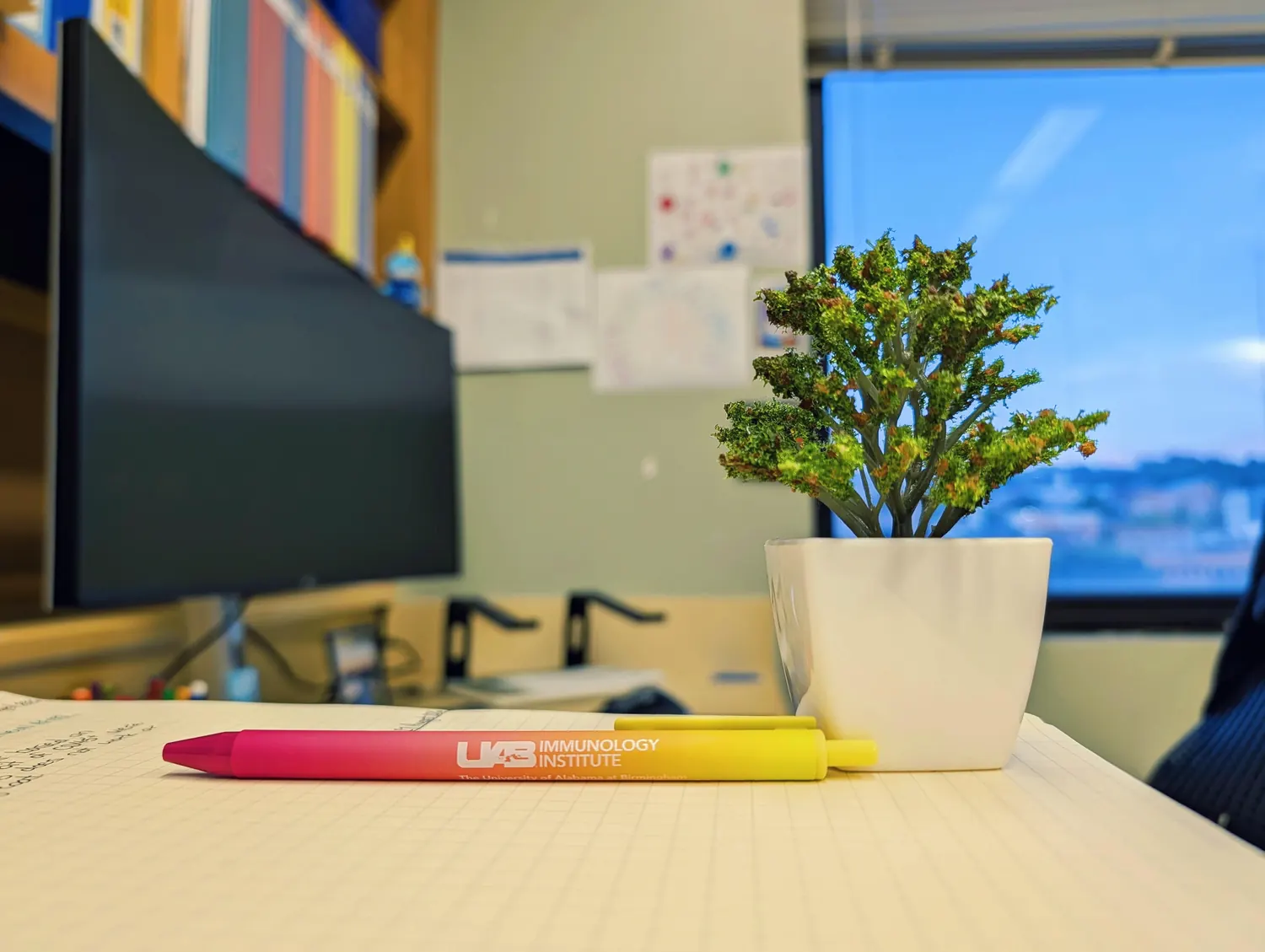 A UAB Immunology Institute pen resting on an open lab notebook beside a monitor and a small plant