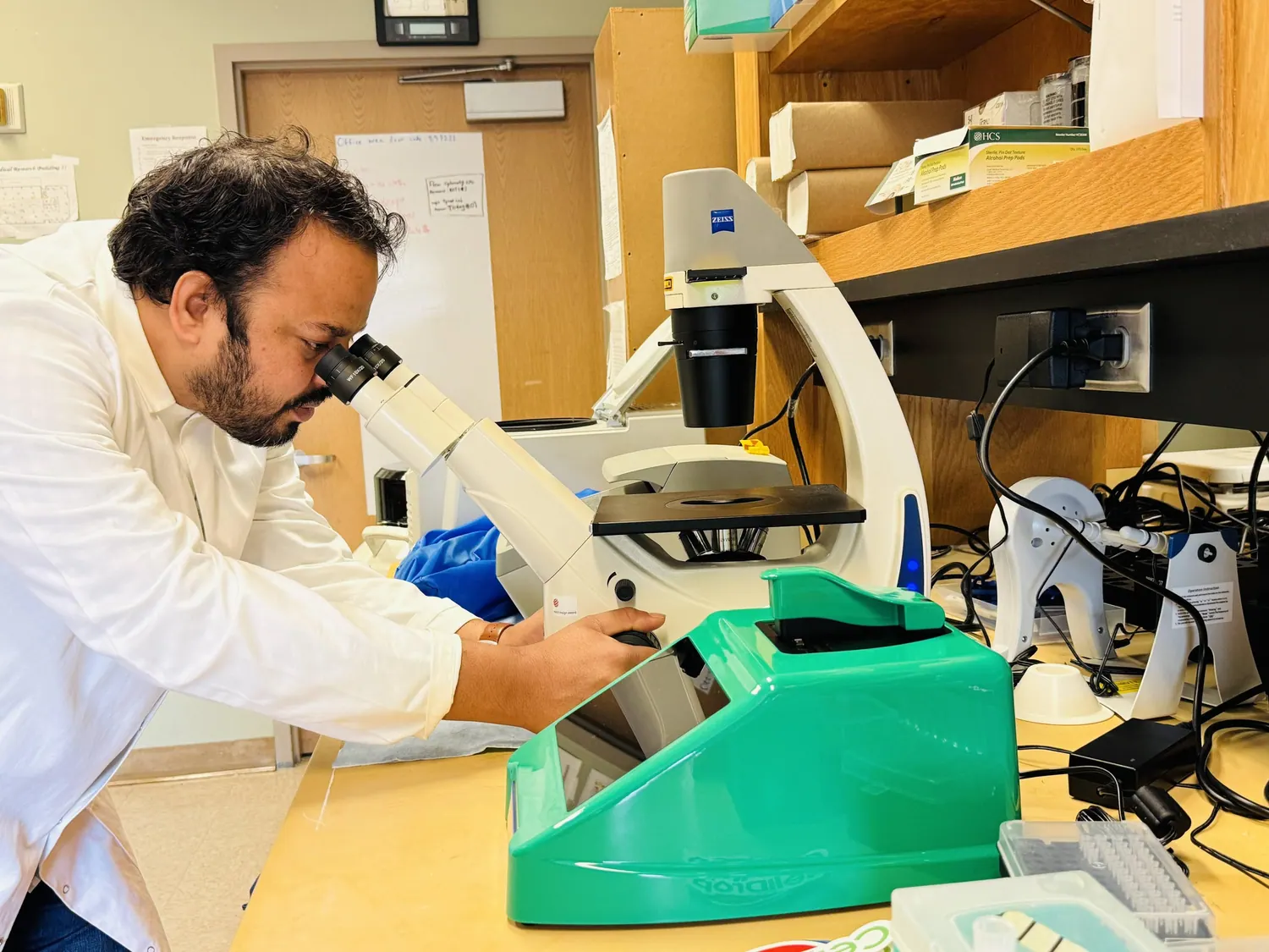 Md Akkas Ali in a lab coat examining a sample down a Zeiss microscope at the bench